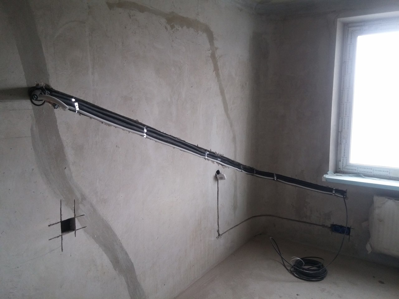 Montaż instalacji klimatyzacyjnej: czarne rury prowadzone wzdłuż surowej ściany, przymocowane metalowymi uchwytami. Widoczne otwory w ścianie pod gniazdka i kable.