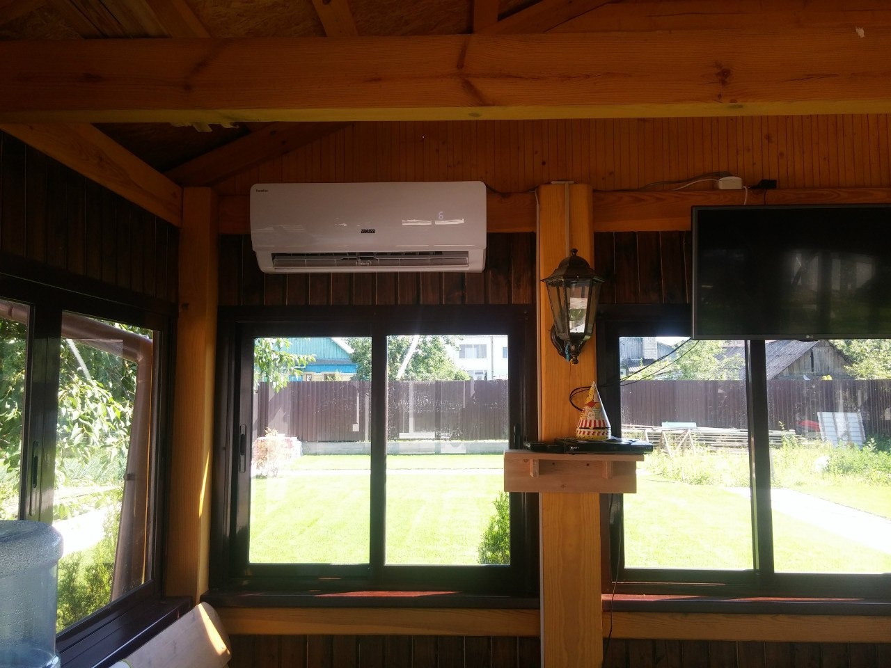 Klimatyzator ścienny Zamussi w drewnianej altanie we Wrocławiu. Widok na ogród przez okna. Obok lampa na ścianie i telewizor. Montaż klimatyzacji w domku letniskowym.