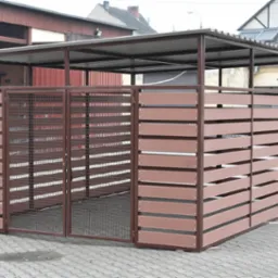 Zadaszona wiata śmietnikowa z brązowymi, poziomymi panelami i metalową siatką, stojąca na szarym bruku. W tle budynki mieszkalne i gospodarcze.