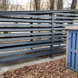 SHIPSTEEL MATEUSZ SKOPIŃSKI - Szara, drewniana konstrukcja osłonowa na śmietniki z poziomymi listwami, na tle bloku mieszkalnego i drzew bez liści, jesienne liście na ziemi.