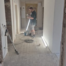 SHIPSTEEL MATEUSZ SKOPIŃSKI - Remont korytarza: mężczyzna szpachluje ścianę, widoczne narzędzia, drabina, podłoga drewniana przed wykończeniem. Oświetlenie robocze. Fragment odsłoniętej cegły.