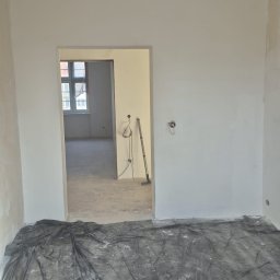 SHIPSTEEL MATEUSZ SKOPIŃSKI - Wnętrze po malowaniu: świeżo pomalowane ściany na biało, podłoga zabezpieczona folią malarską. Widoczne narzędzia malarskie w przejściu do kolejnego pomieszczenia.