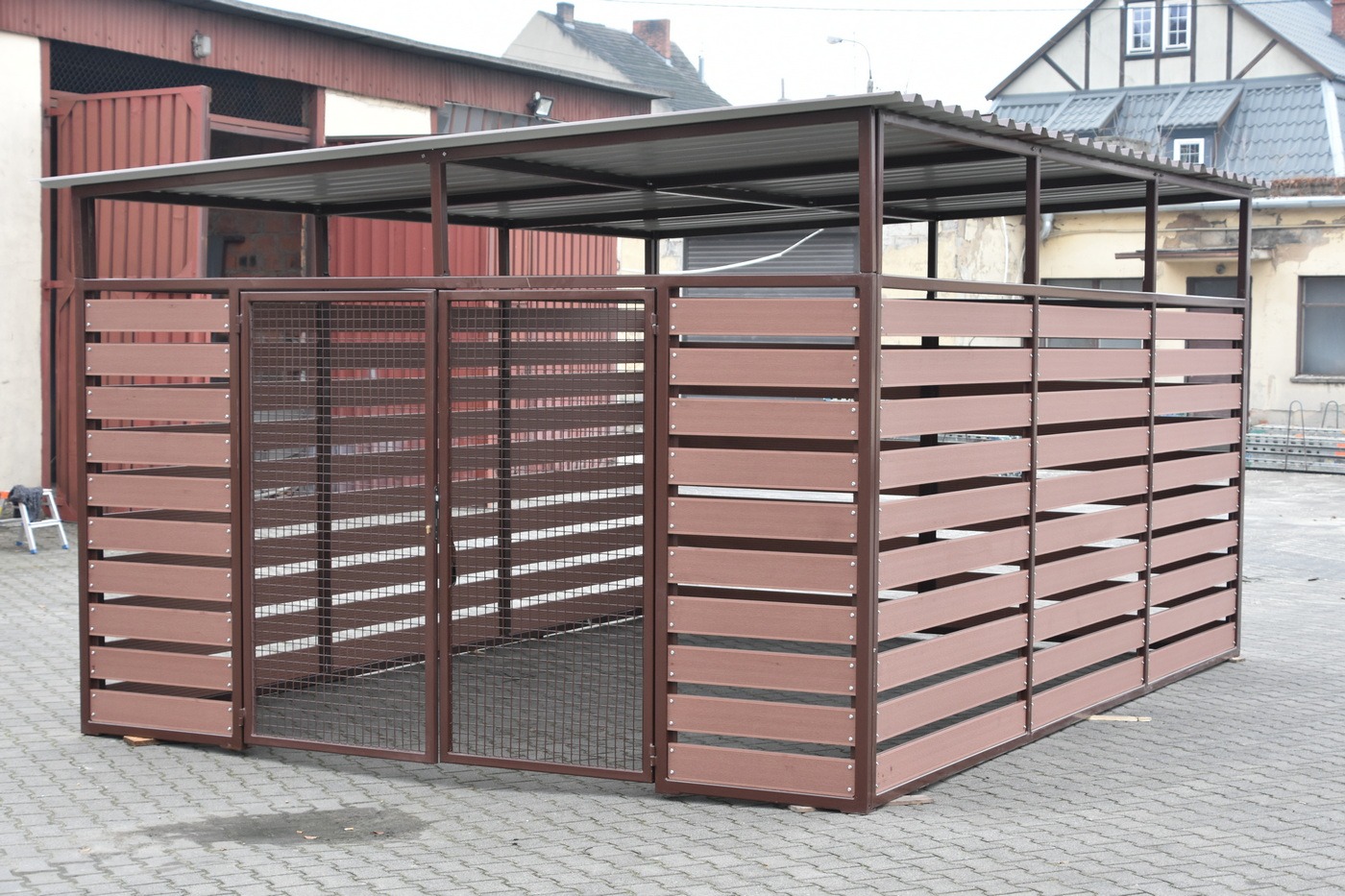 Zadaszona wiata śmietnikowa z brązowymi, poziomymi panelami i metalową siatką, stojąca na szarym bruku. W tle budynki mieszkalne i gospodarcze.
