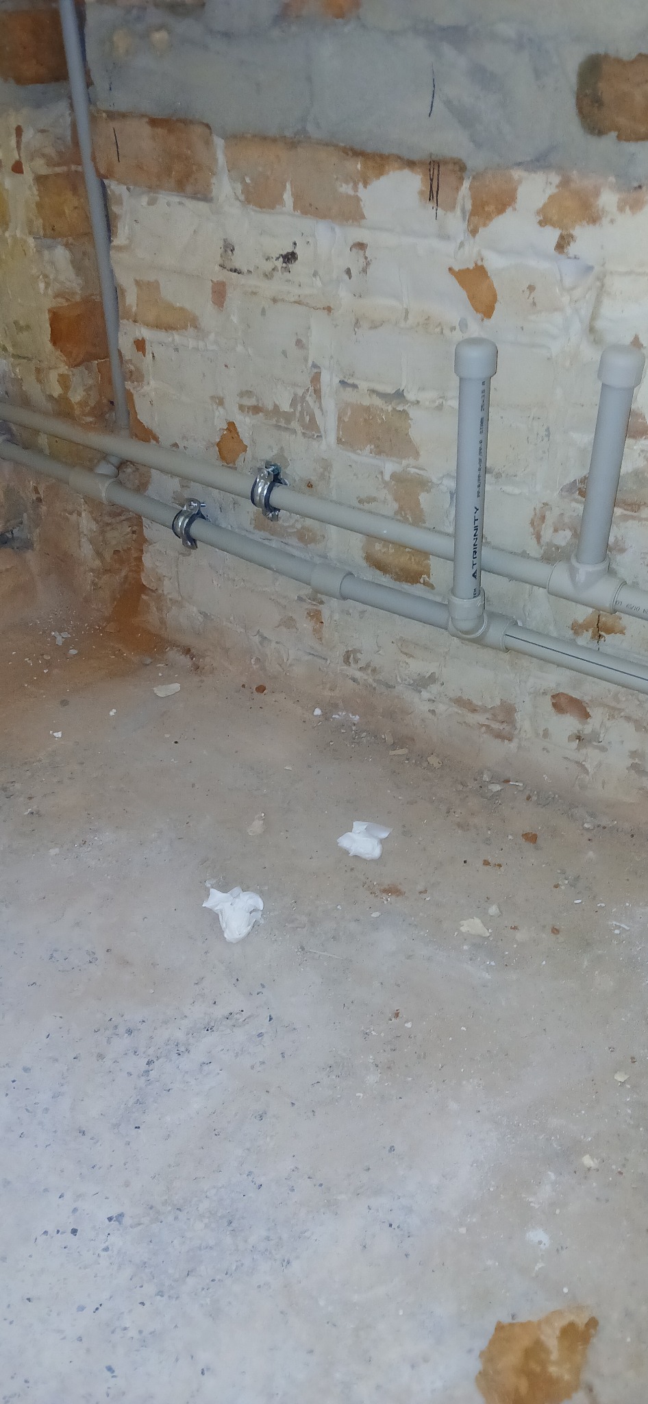 Szare rury instalacji wodnej zamocowane do ściany z cegły. Dwa uchwyty mocujące. Na podłodze widoczne zabrudzenia i resztki materiałów.
