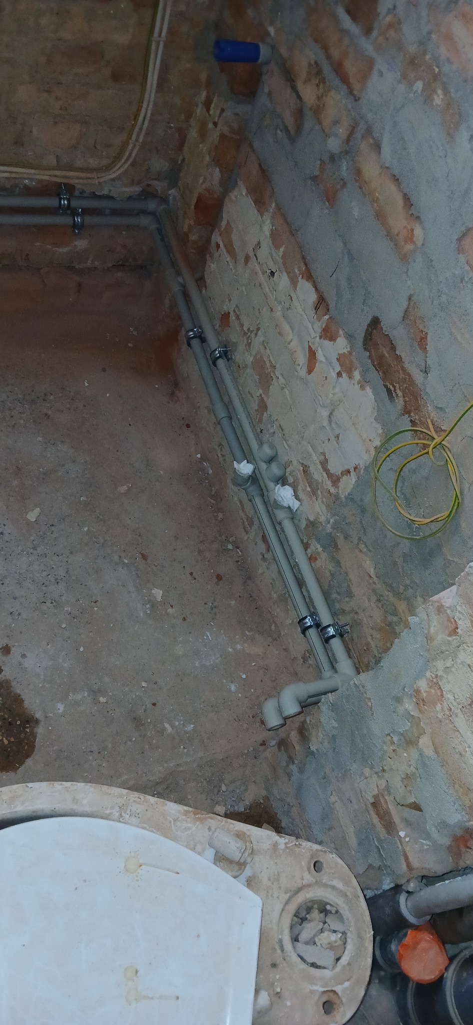 Szare rury instalacji wodnej zamocowane do ceglanej ściany z widoczną muszlą klozetową w trakcie prac remontowych. Żółty kabel elektryczny w tle.