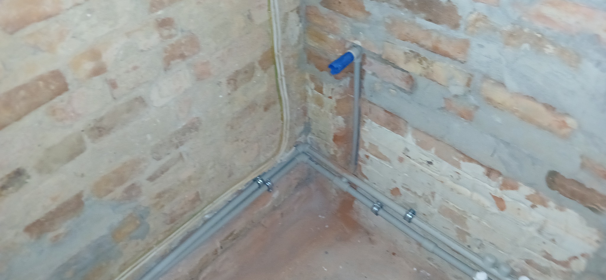 Fragment ściany z cegły z poprowadzonymi rurami instalacji wodnej, zabezpieczonymi metalowymi uchwytami. Widoczny fragment podłogi i pionowa rura z niebieskim zakończeniem.