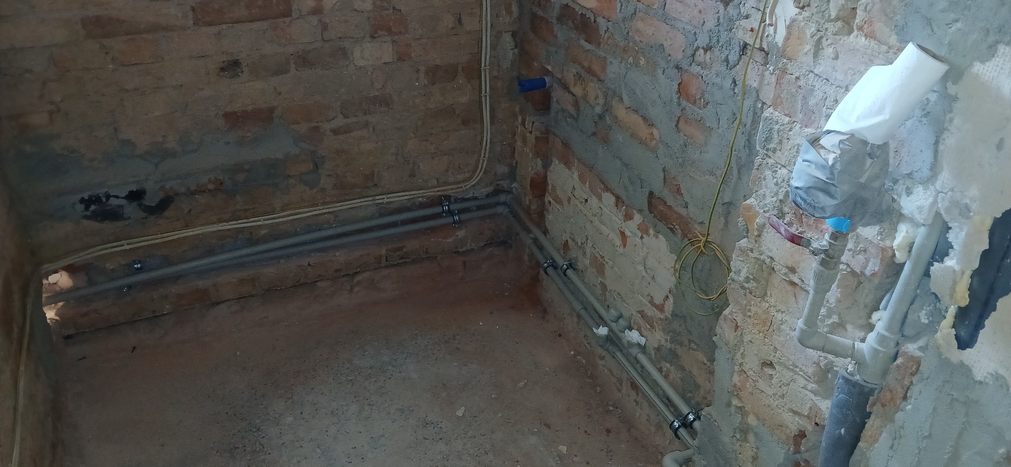 Szare rury instalacji wodnej zamontowane na ceglanej ścianie z widocznymi śladami zaprawy. Fragment pomieszczenia w trakcie prac instalacyjnych.
