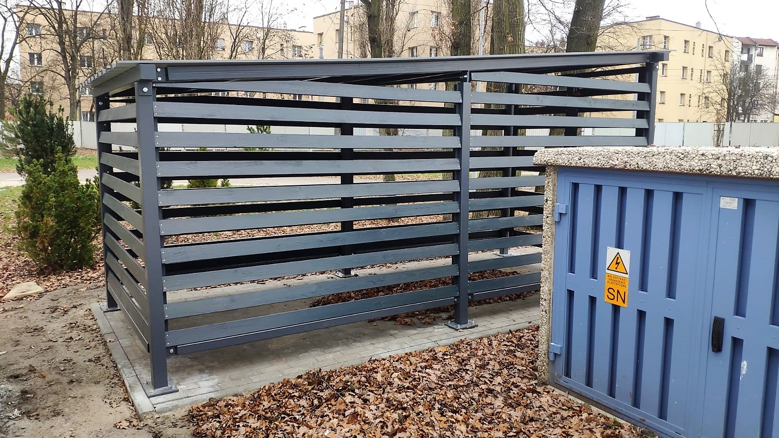 Szara, drewniana konstrukcja osłonowa na śmietniki z poziomymi listwami, na tle bloku mieszkalnego i drzew bez liści, jesienne liście na ziemi.