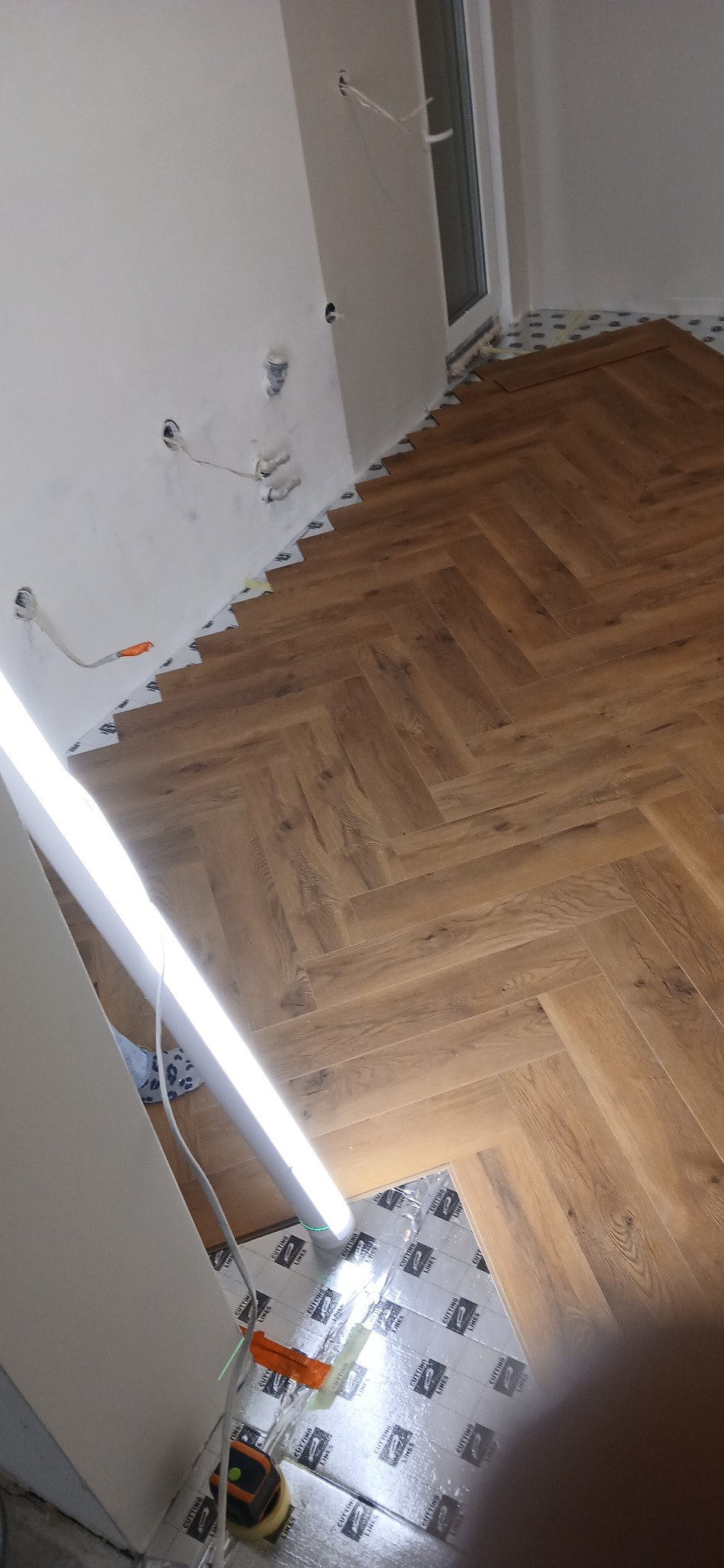 Układanie paneli podłogowych w jodełkę francuską, widoczne narzędzia i podkład izolacyjny. Częściowo wykończone wnętrze z widocznymi przewodami elektrycznymi.