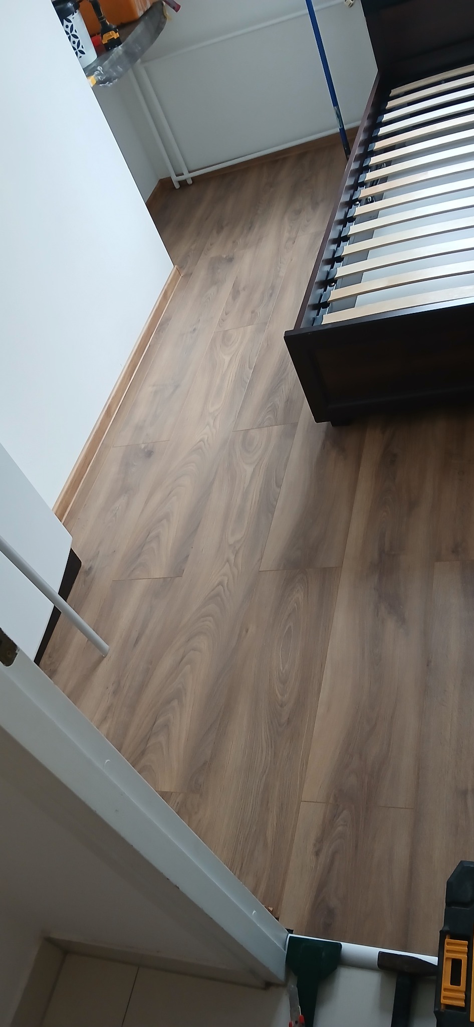 Ułożona podłoga z paneli drewnopodobnych w jasnym odcieniu, widoczna część ramy łóżka i biała ściana z listwą przypodłogową. Widoczne narzędzia.