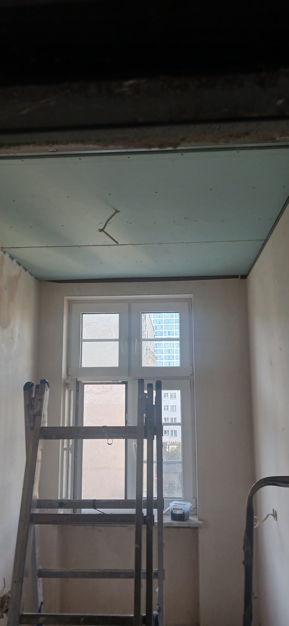 Montaż sufitu podwieszanego z płyt gipsowo-kartonowych w trakcie prac wykończeniowych. Widoczna drabina, okno i wystające kable. Ściany przygotowane do malowania.