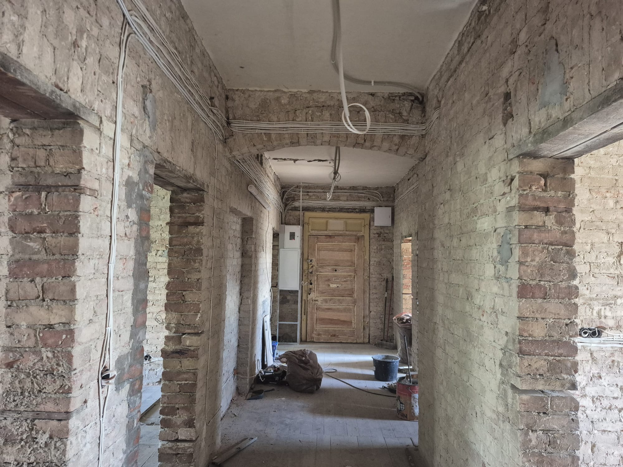 Korytarz w trakcie remontu z odsłoniętą cegłą, widoczne przewody elektryczne i narzędzia. Charakterystyczne łukowe przejście. Oświetlenie naturalne.