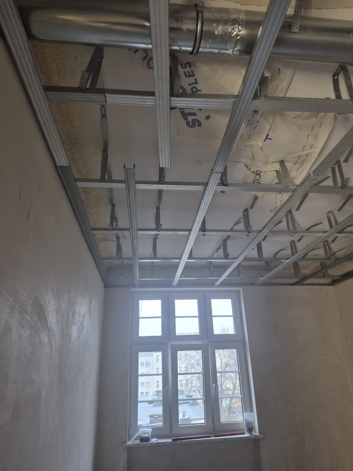 Montaż metalowej konstrukcji sufitu podwieszanego z widocznymi elementami izolacji i rurami wentylacyjnymi, w tle okno. Etap prac wykończeniowych.