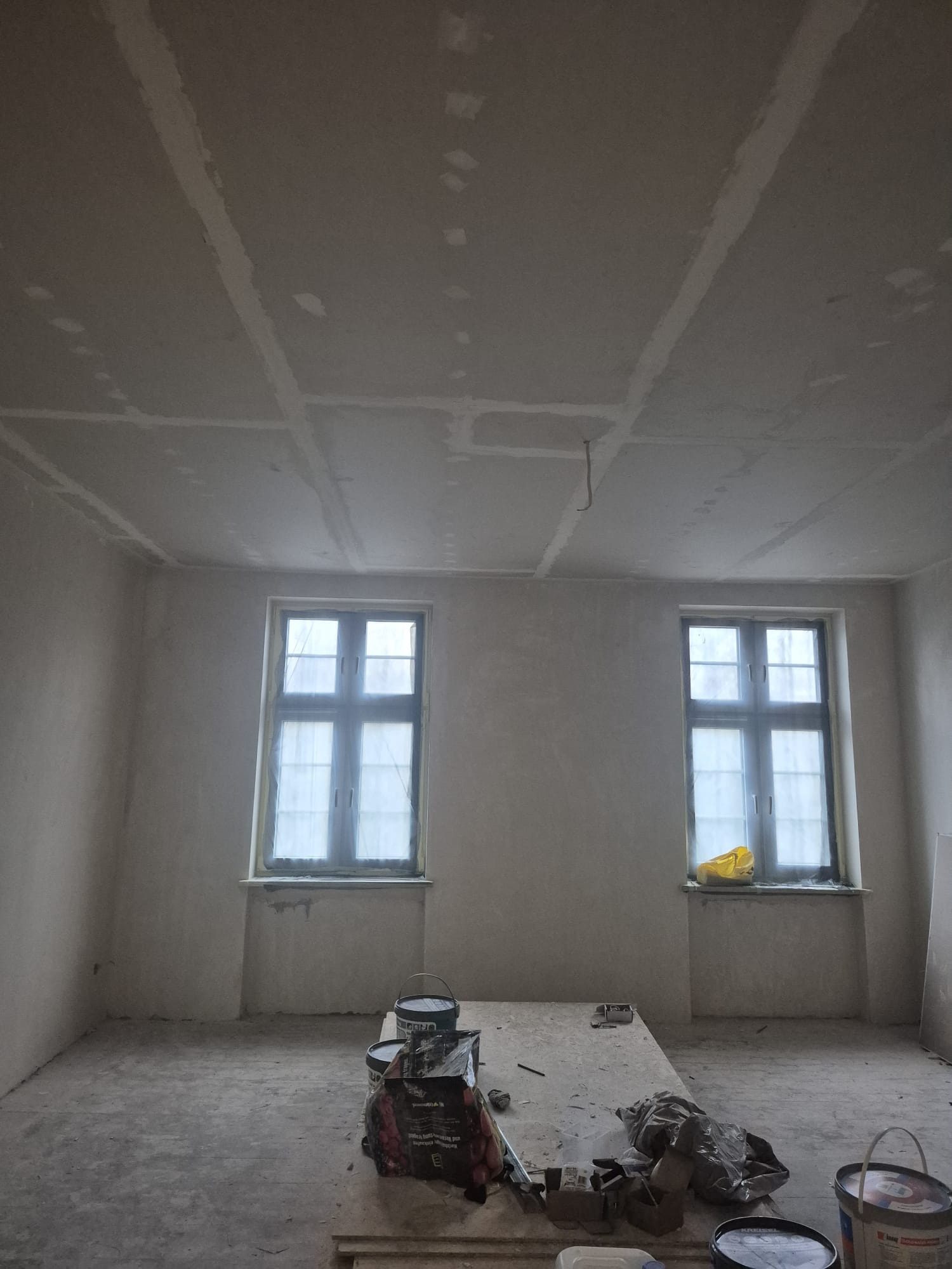 Pokój w trakcie remontu: ściany i sufit po szpachlowaniu, widoczne łączenia płyt gipsowych, okna zabezpieczone folią, narzędzia i materiały malarskie na podłodze.