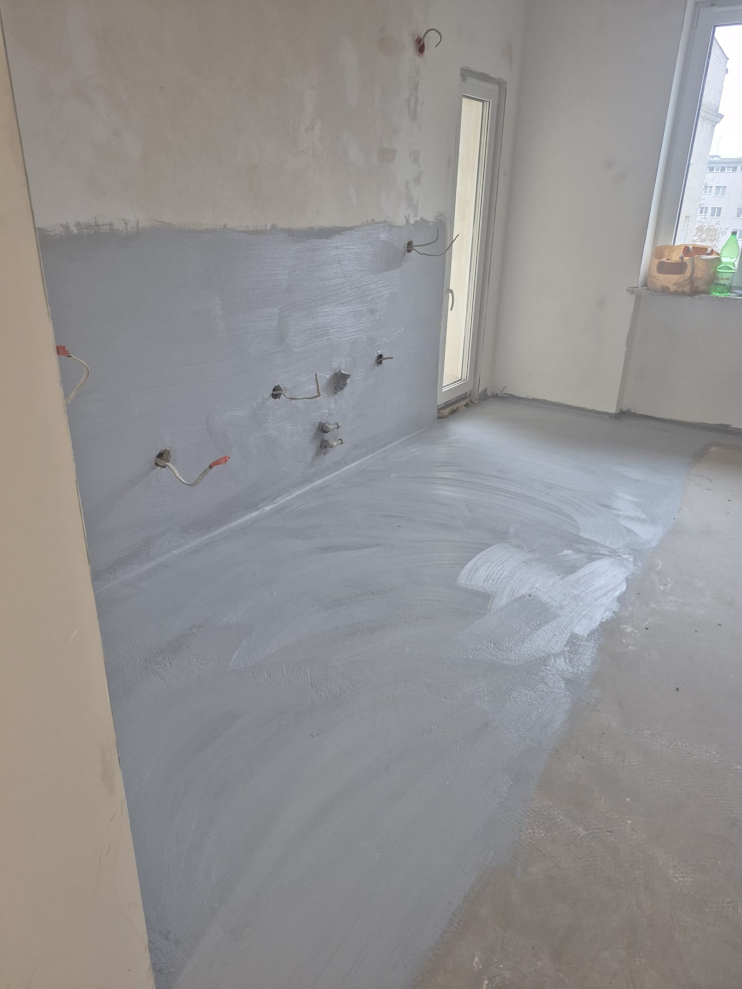 Szara, świeżo pomalowana ściana i podłoga w pustym pokoju z oknem. Widoczne wystające kable i rury. Przygotowanie do dalszych prac wykończeniowych.