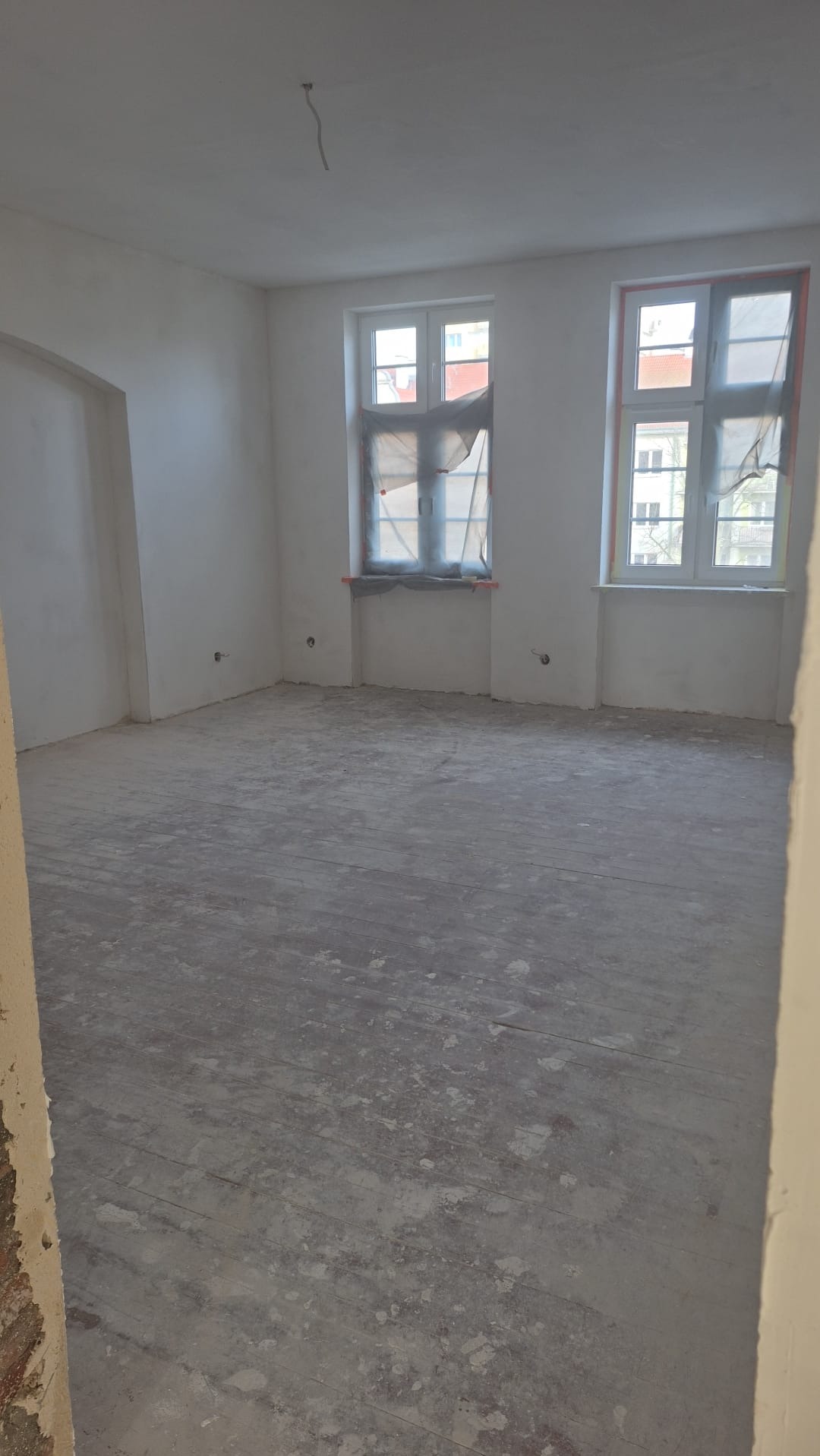 Wnętrze pokoju w trakcie remontu. Widoczne dwa okna z zabezpieczeniami, surowe ściany i podłoga z desek pokryta pyłem. Fragment ściany z cegły w lewym dolnym rogu.