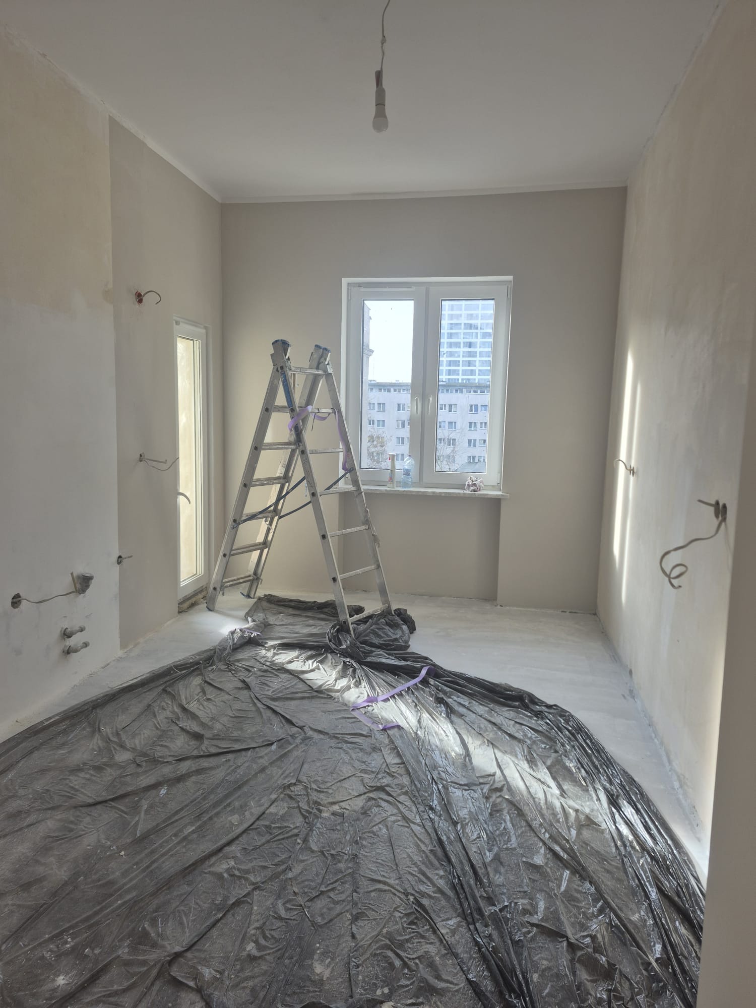 Pokój w trakcie przygotowania do malowania: zabezpieczona folią podłoga, drabina oparta o ścianę, ściany częściowo zagruntowane, widoczne wyprowadzenia instalacji elektrycznej.