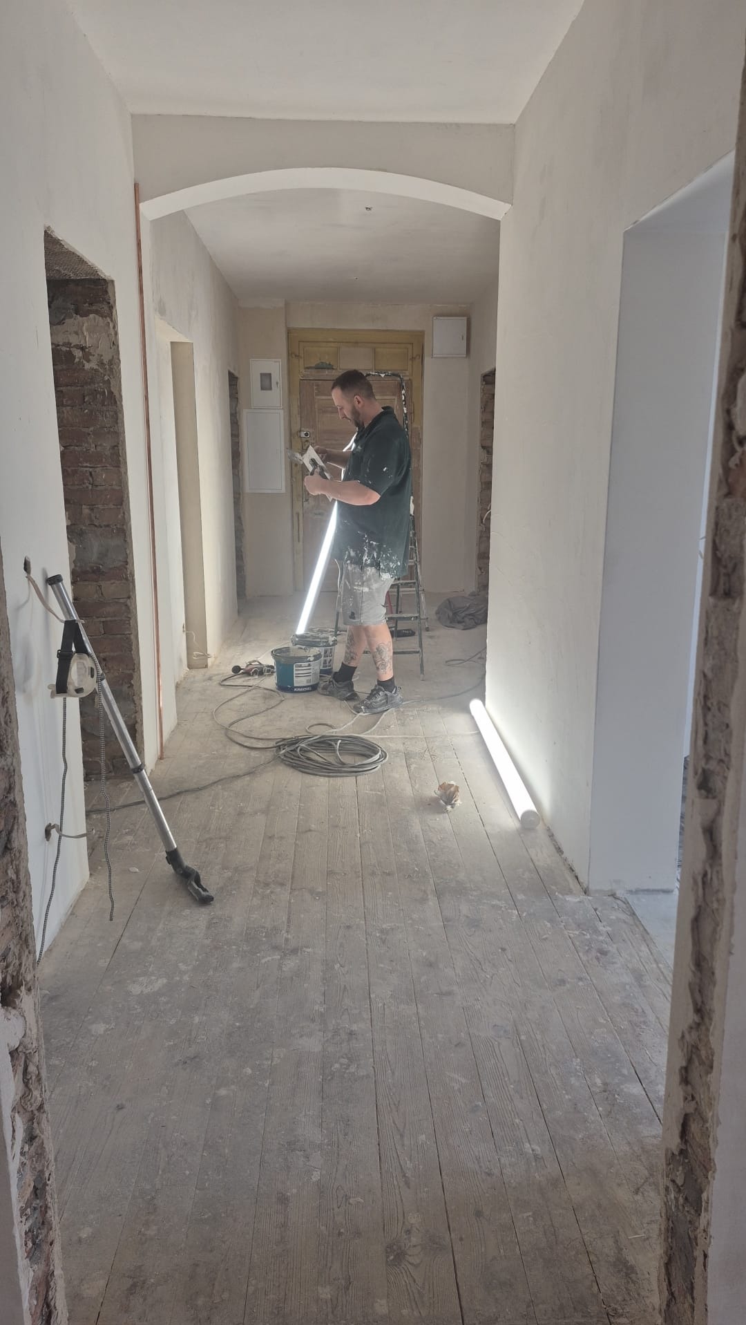 Remont korytarza: mężczyzna szpachluje ścianę, widoczne narzędzia, drabina, podłoga drewniana przed wykończeniem. Oświetlenie robocze. Fragment odsłoniętej cegły.
