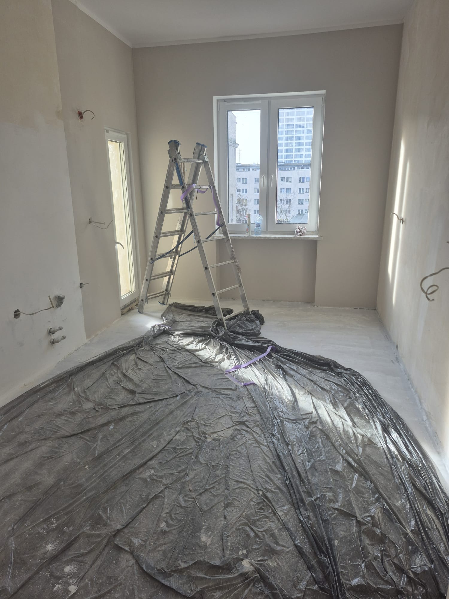 Pokój w trakcie malowania: zabezpieczona podłoga folią, drabina oparta o ścianę, widoczne puszki elektryczne. Przygotowanie do malowania ścian na jasny odcień.