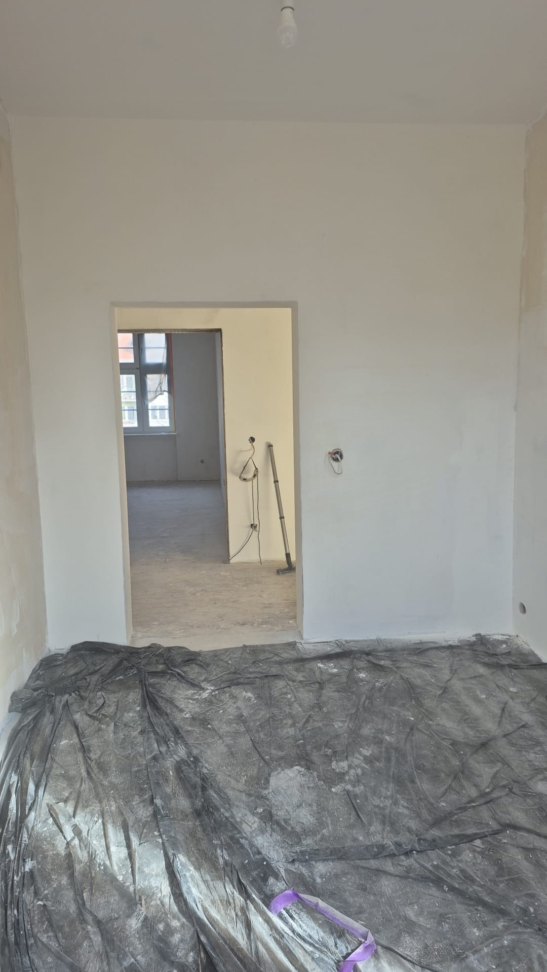Wnętrze po malowaniu: świeżo pomalowane ściany na biało, podłoga zabezpieczona folią malarską. Widoczne narzędzia malarskie w przejściu do kolejnego pomieszczenia.