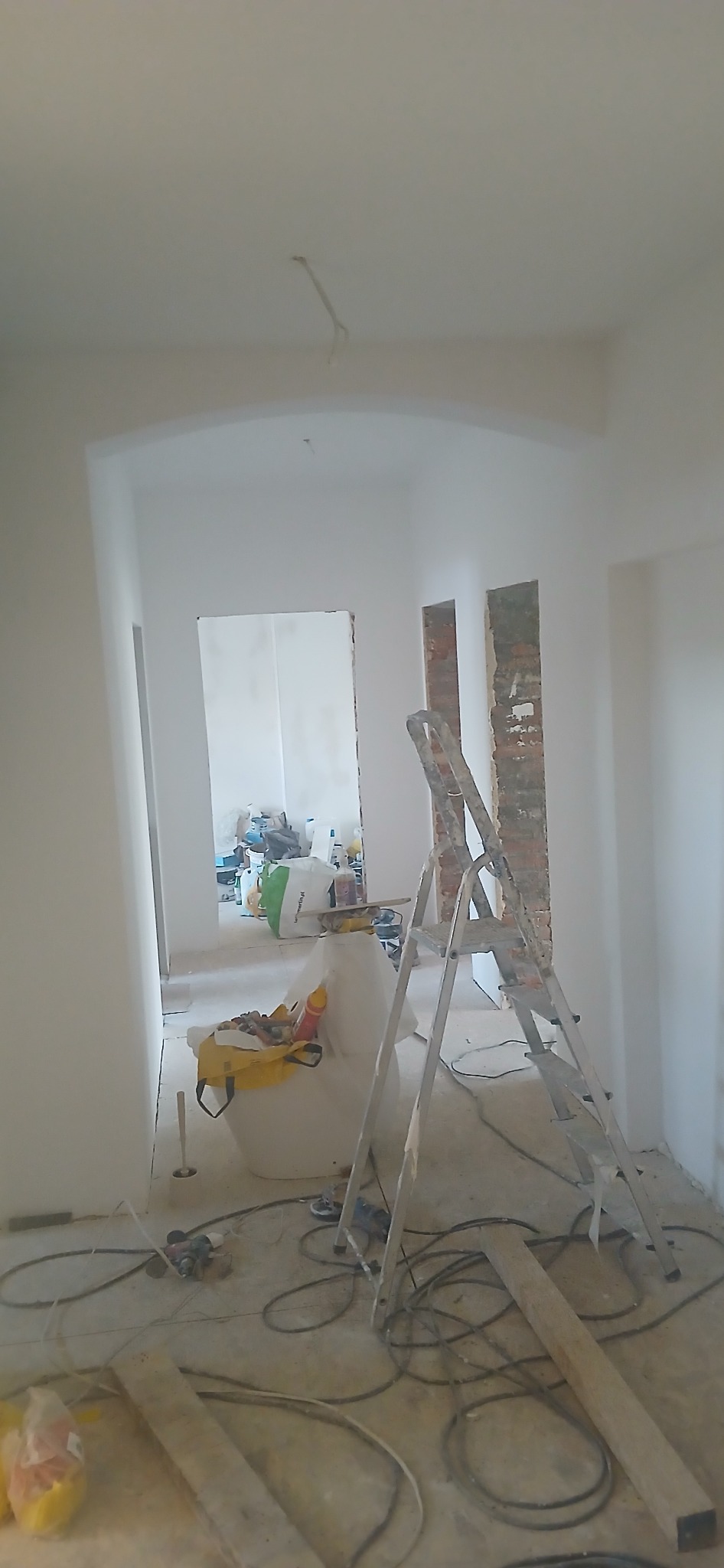 Wnętrze w trakcie malowania: białe ściany, drabina oparta o ścianę z odsłoniętą cegłą, narzędzia malarskie i kable na podłodze. Widok na kolejne pomieszczenie.