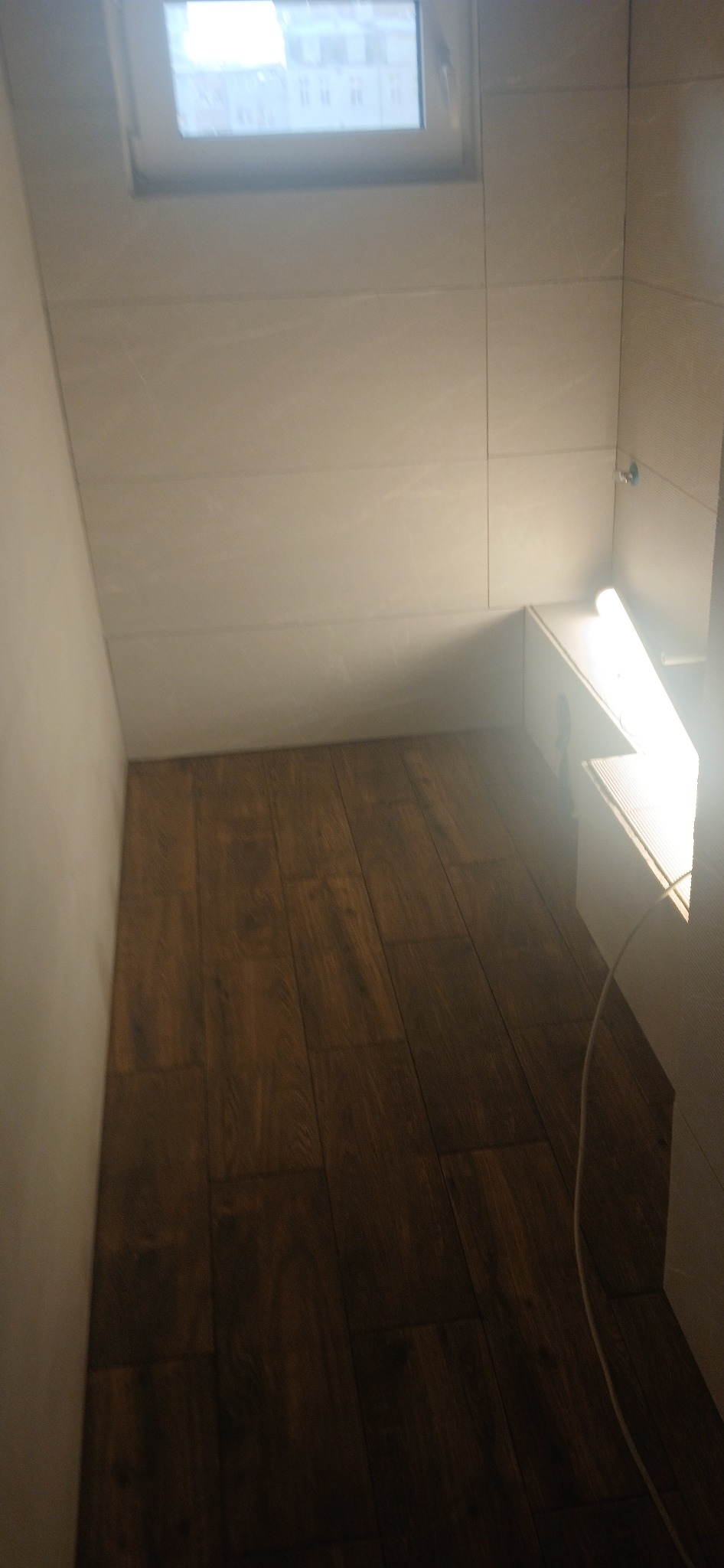 Ułożona podłoga z paneli imitujących drewno w małym pomieszczeniu z oknem i białymi ścianami. Widoczny fragment instalacji elektrycznej.