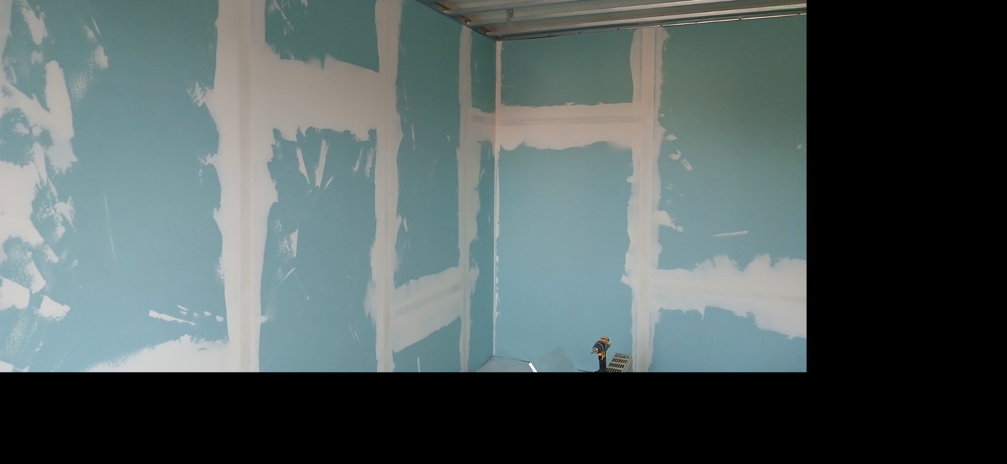 Ściana z płyt gipsowo-kartonowych, wstępnie wyszpachlowana na łączeniach, przygotowana do malowania. Widoczny metalowy profil sufitowy i wkrętarka akumulatorowa.