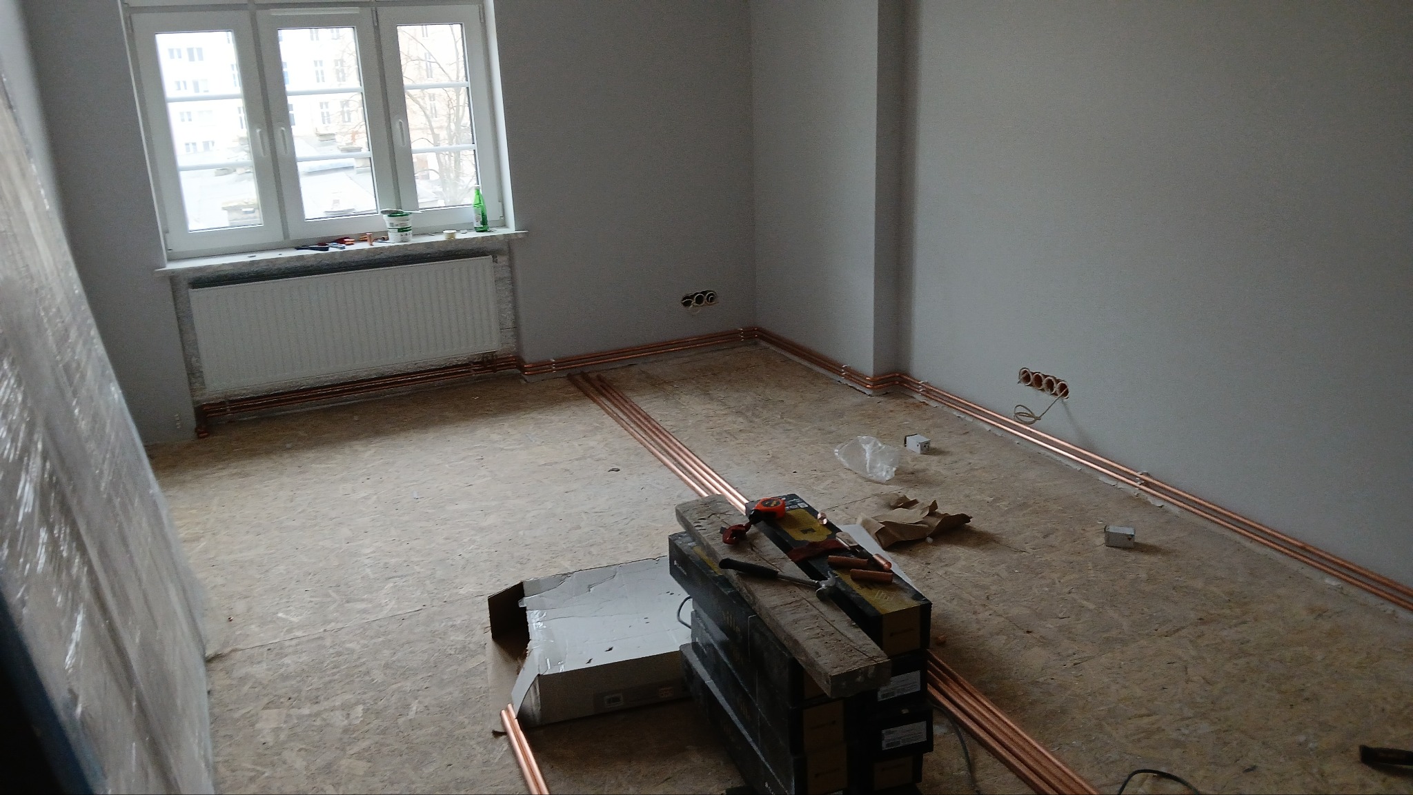 Miedziane rury grzewcze ułożone wzdłuż ścian i pod grzejnikiem w pokoju z surową podłogą. Widoczne narzędzia i materiały budowlane. Etap prac instalacyjnych.