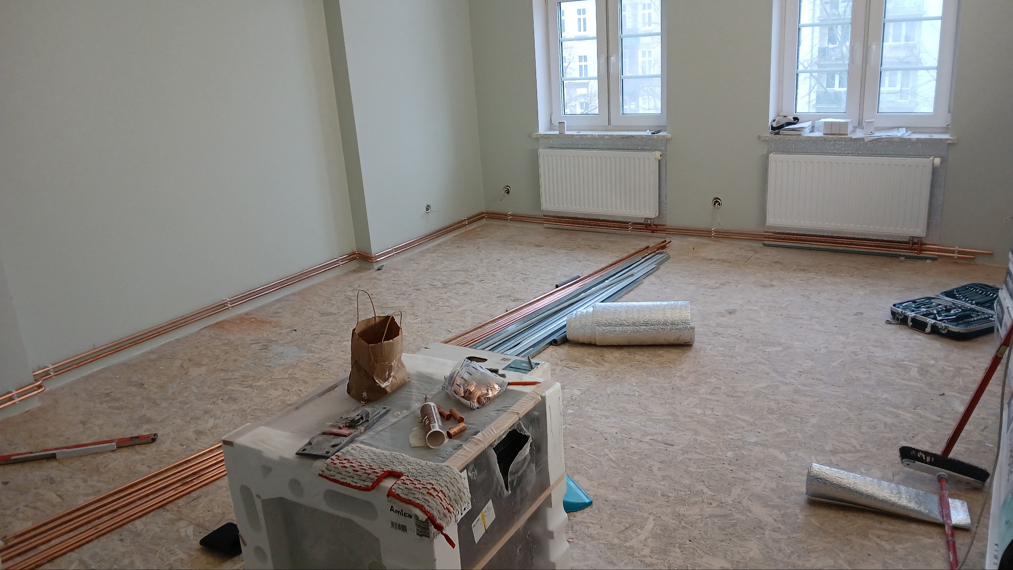 Wnętrze w trakcie instalacji ogrzewania podłogowego: miedziane rury ułożone wzdłuż ścian, narzędzia i materiały na stole, widoczne grzejniki pod oknami. Etap prac.