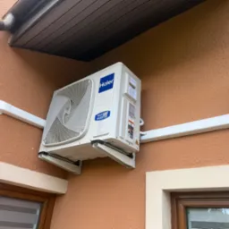 Zamontowana klimatyzacja Haier na zewnątrz budynku o brzoskwiniowej elewacji, widoczna instalacja elektryczna w białej listwie, lampa zewnętrzna i okno.