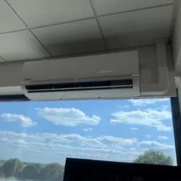 Biała klimatyzacja Kaisai Eco zamontowana pod sufitem, widok zza okna na błękitne niebo i zieleń. Widoczne elementy instalacji.