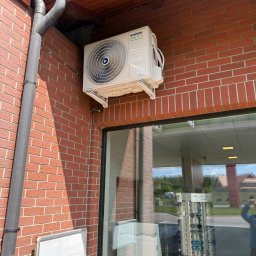 AIR & SUN Hubert Małkiewicz - Klimatyzator KAISAI ECO zamontowany na ceglanej ścianie budynku, widoczny system odprowadzania wody, fragment okna z odbiciem otoczenia.