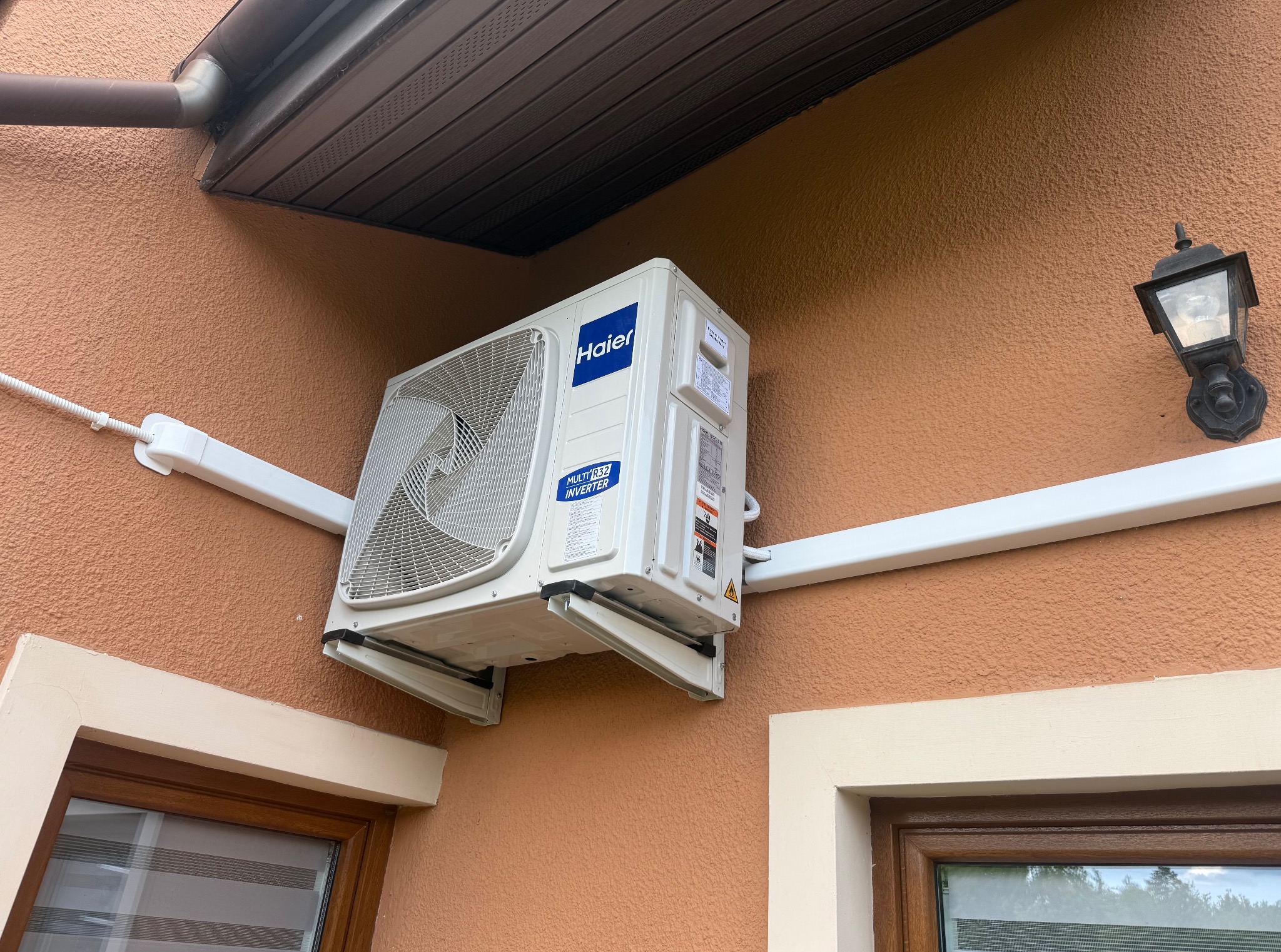 Zamontowana klimatyzacja Haier na zewnątrz budynku o brzoskwiniowej elewacji, widoczna instalacja elektryczna w białej listwie, lampa zewnętrzna i okno.
