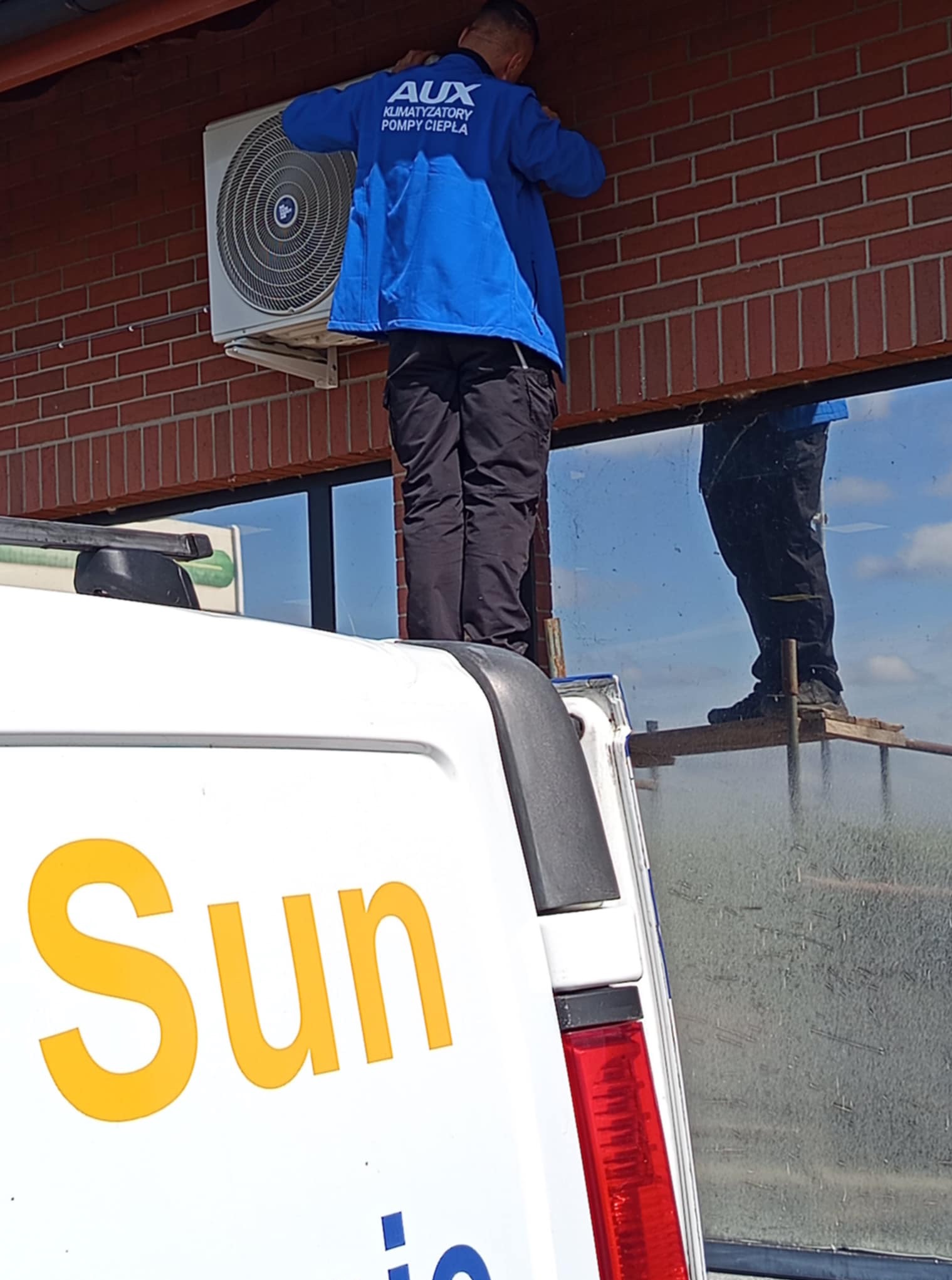 Montaż klimatyzacji AUX na ceglanej ścianie budynku. Instalator w niebieskiej kurtce firmowej stoi na dachu białego samochodu dostawczego.