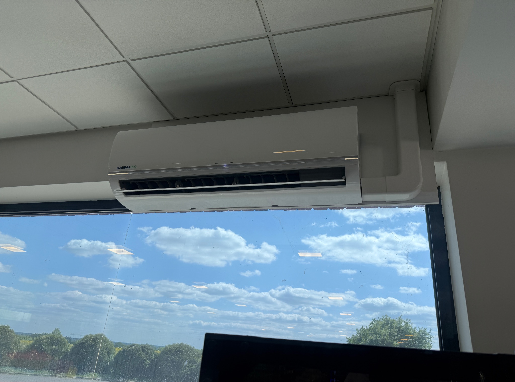 Biała klimatyzacja Kaisai Eco zamontowana pod sufitem, widok zza okna na błękitne niebo i zieleń. Widoczne elementy instalacji.
