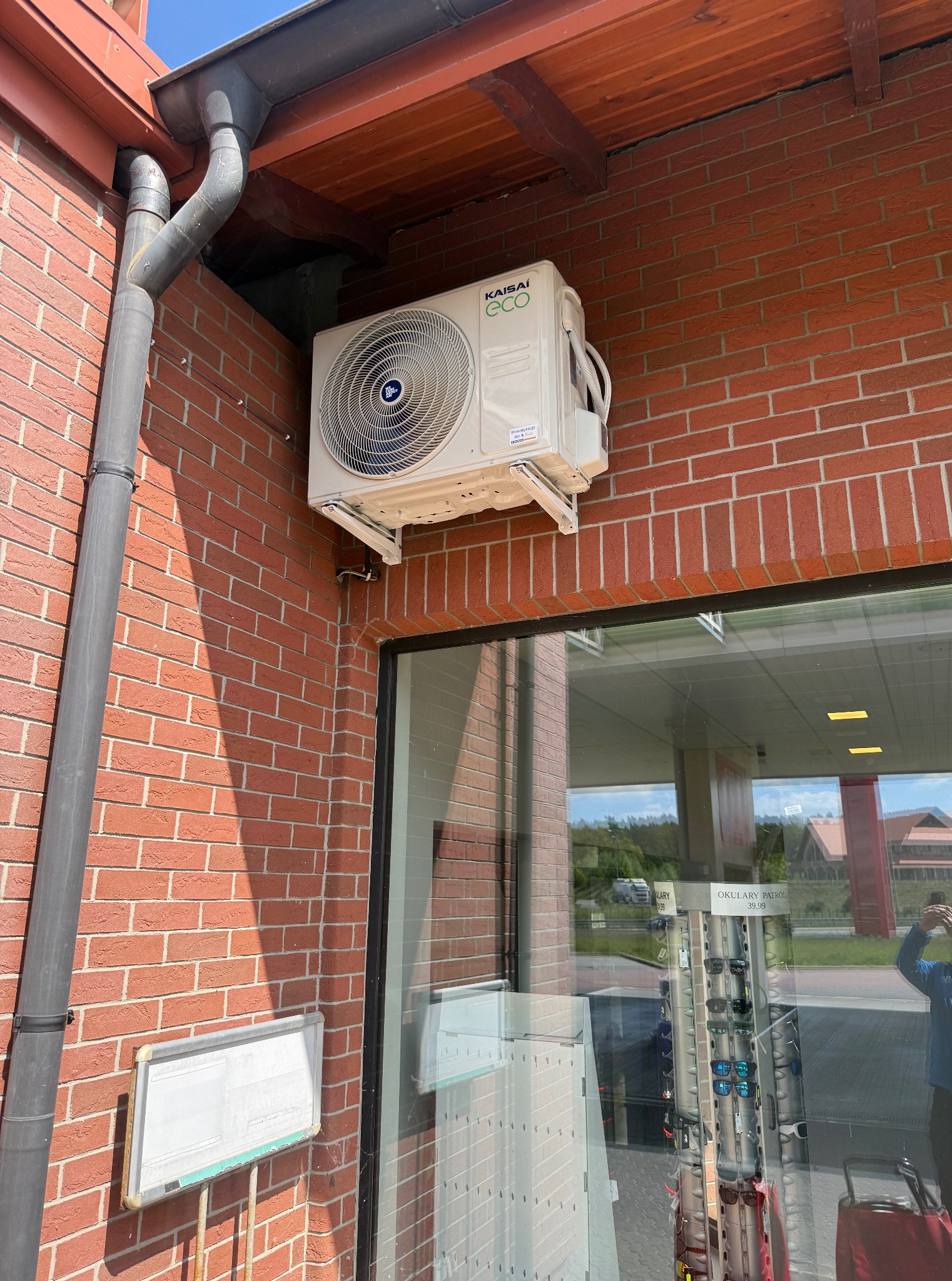 Klimatyzator KAISAI ECO zamontowany na ceglanej ścianie budynku, widoczny system odprowadzania wody, fragment okna z odbiciem otoczenia.