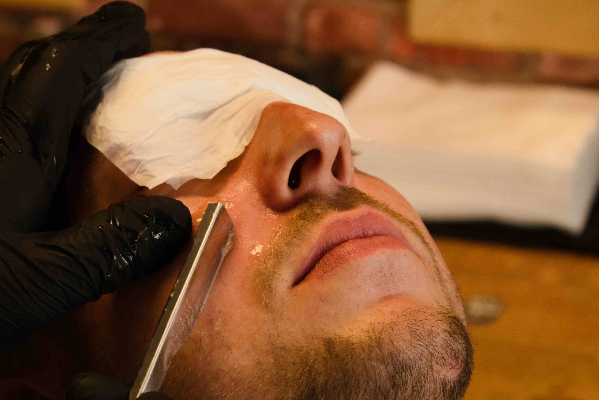 Golenie brzytwą brody mężczyzny z ręcznikiem na twarzy. Ręka w czarnej rękawiczce trzyma ostrze. Widoczny zarost i piana do golenia. Ujęcie z bliska, perspektywa od dołu.