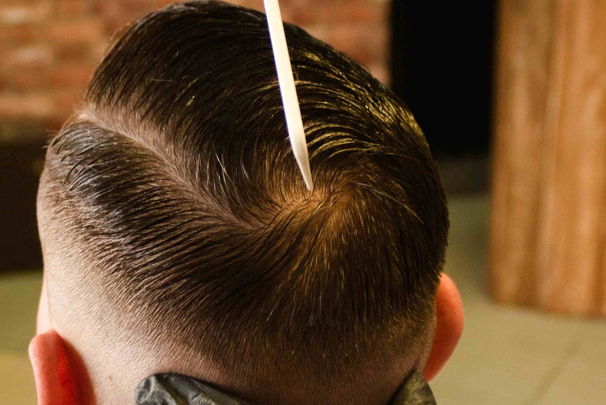 Męska fryzura z wyraźnym przedziałkiem, utrwalona pomadą, z bliska. Włosy zaczesane do tyłu, widoczny grzebień wbijający się w przedziałek. Krótkie boki.
