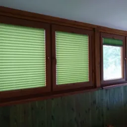 Drewniane okna z zielonymi plisowanymi roletami, częściowo zasłoniętymi, w pomieszczeniu z boazerią. Widok z wnętrza na zewnątrz, z oknami w ciemnej ramie.