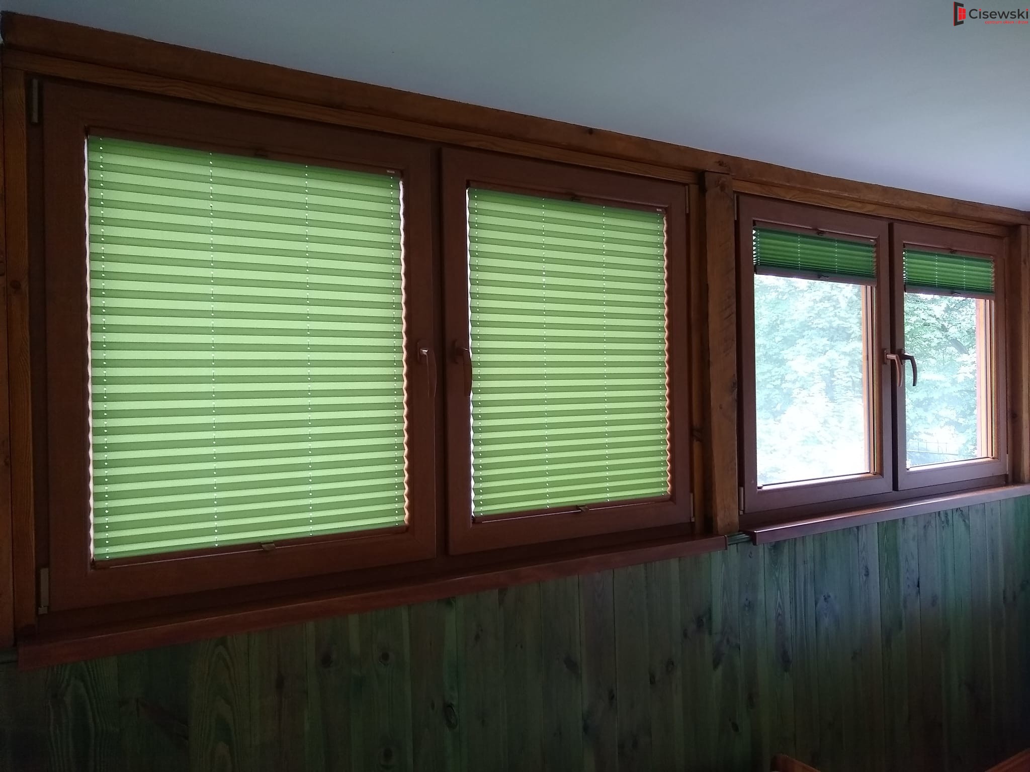 Drewniane okna z zielonymi plisowanymi roletami, częściowo zasłoniętymi, w pomieszczeniu z boazerią. Widok z wnętrza na zewnątrz, z oknami w ciemnej ramie.