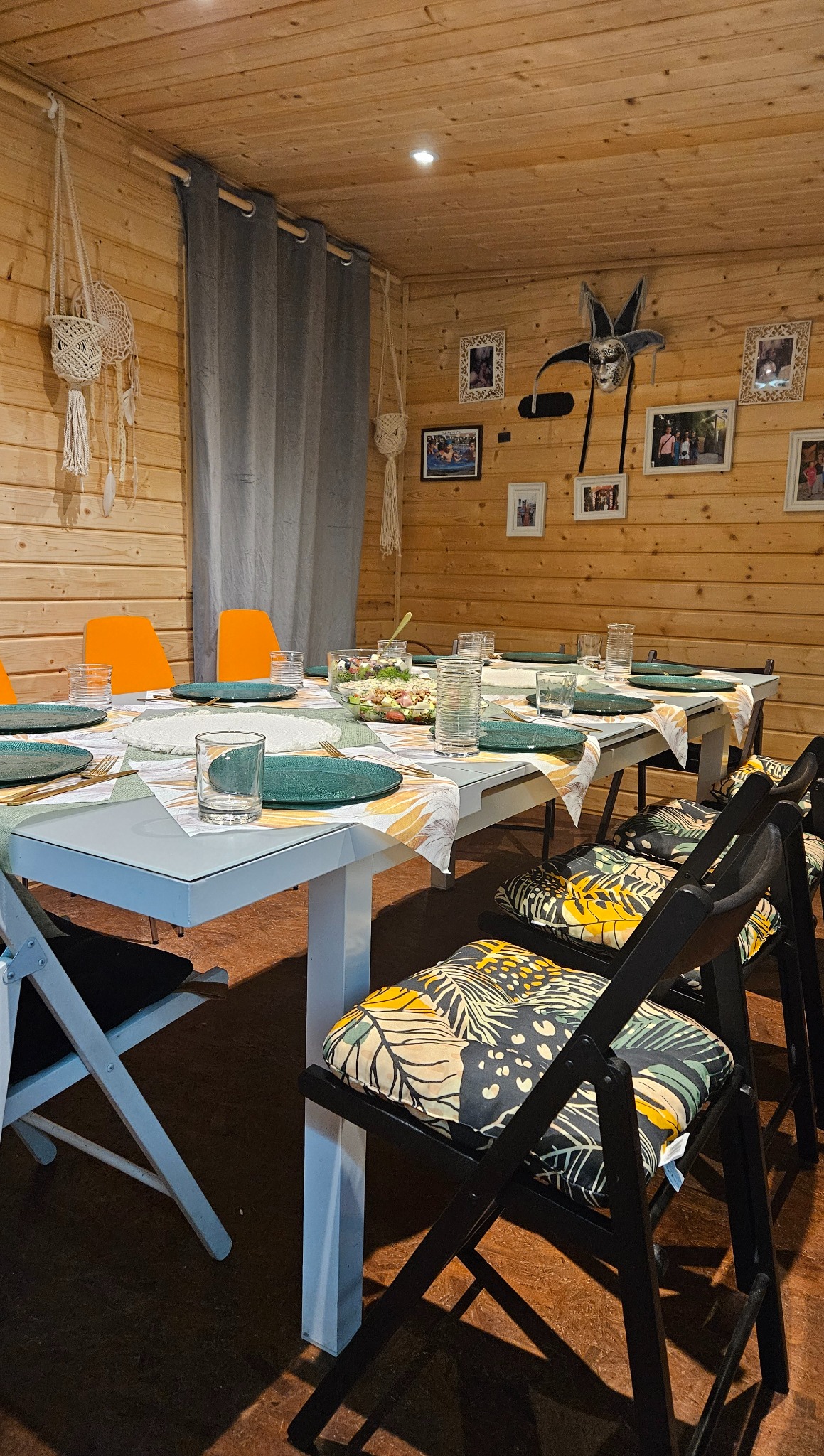 Drewniane wnętrze jadalni z nakrytym stołem, talerzami w kolorze butelkowej zieleni, szklankami i dekoracyjnymi krzesłami z motywem liści. Przytulna aranżacja.