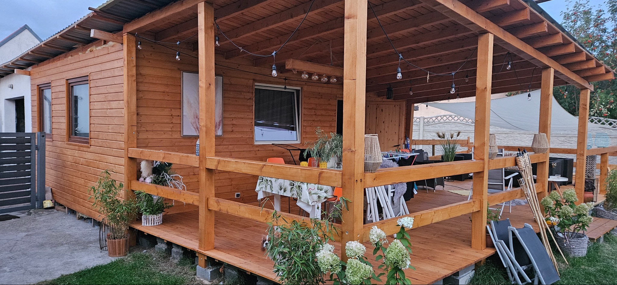 Drewniany dom szkieletowy z zadaszonym tarasem, dekoracje świetlne, rośliny w donicach i meble ogrodowe tworzą przytulną przestrzeń relaksu.