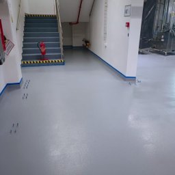 Arte Chemia Budowlana Sp. z o.o. - Jasnoszara, gładka podłoga w hali produkcyjnej, zabezpieczona niebieską taśmą przy ścianach i schodach. Widoczne schody z pasami ostrzegawczymi i gaśnica.