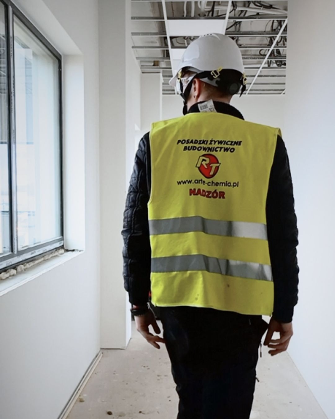 Nadzór budowlany w kamizelce z napisem 'Posadzki żywiczne budownictwo' idzie korytarzem z oknem. Widoczny surowy stan wykończenia wnętrza.