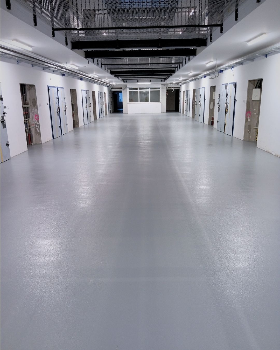 Długa hala z szarą, gładką posadzką. Widoczne drzwi po bokach i metalowa konstrukcja pod sufitem. Jednolite, jasne oświetlenie podkreśla równą powierzchnię.