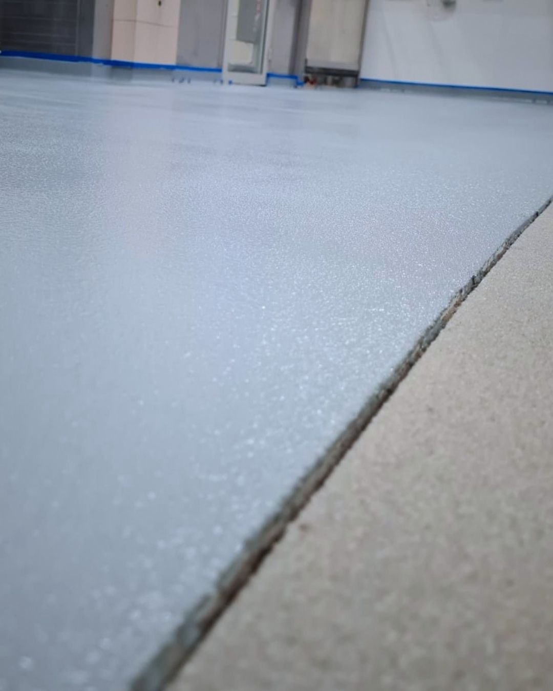 Jasnoszara, gładka posadzka żywiczna w pomieszczeniu, widoczna szczelina dylatacyjna i niebieska taśma malarska przy ścianie. Minimalistyczny design.