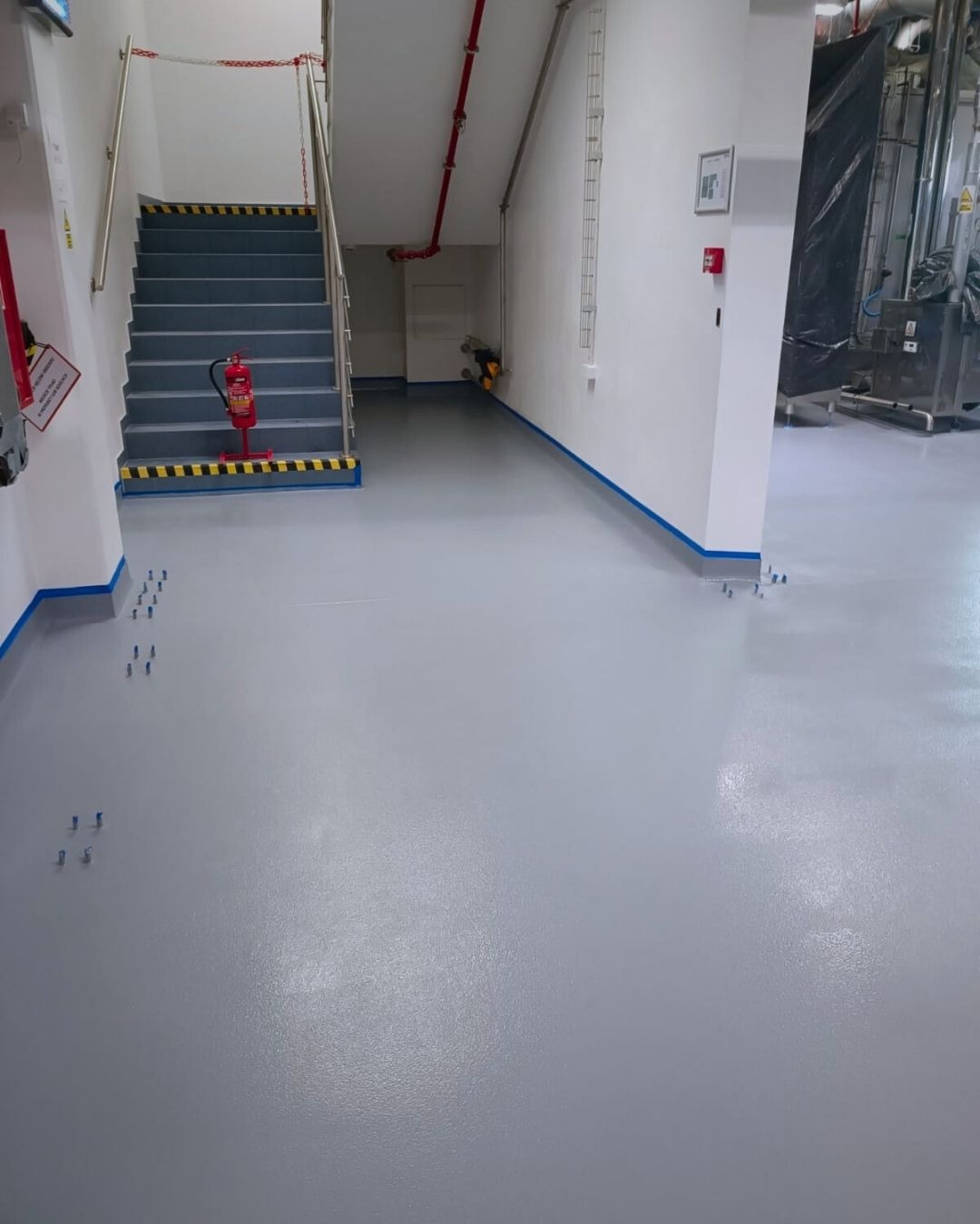 Jasnoszara, gładka podłoga w hali produkcyjnej, zabezpieczona niebieską taśmą przy ścianach i schodach. Widoczne schody z pasami ostrzegawczymi i gaśnica.