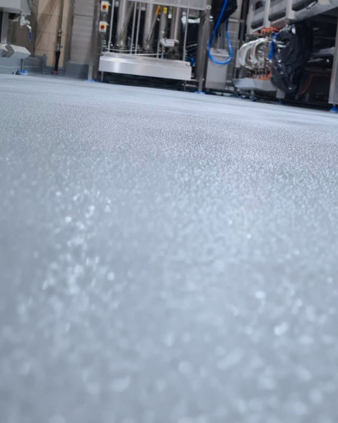 Jasnoszara, gładka posadzka w hali produkcyjnej z maszynami w tle. Widoczna struktura powierzchni, jednolite wykończenie, higieniczna przestrzeń przemysłowa.