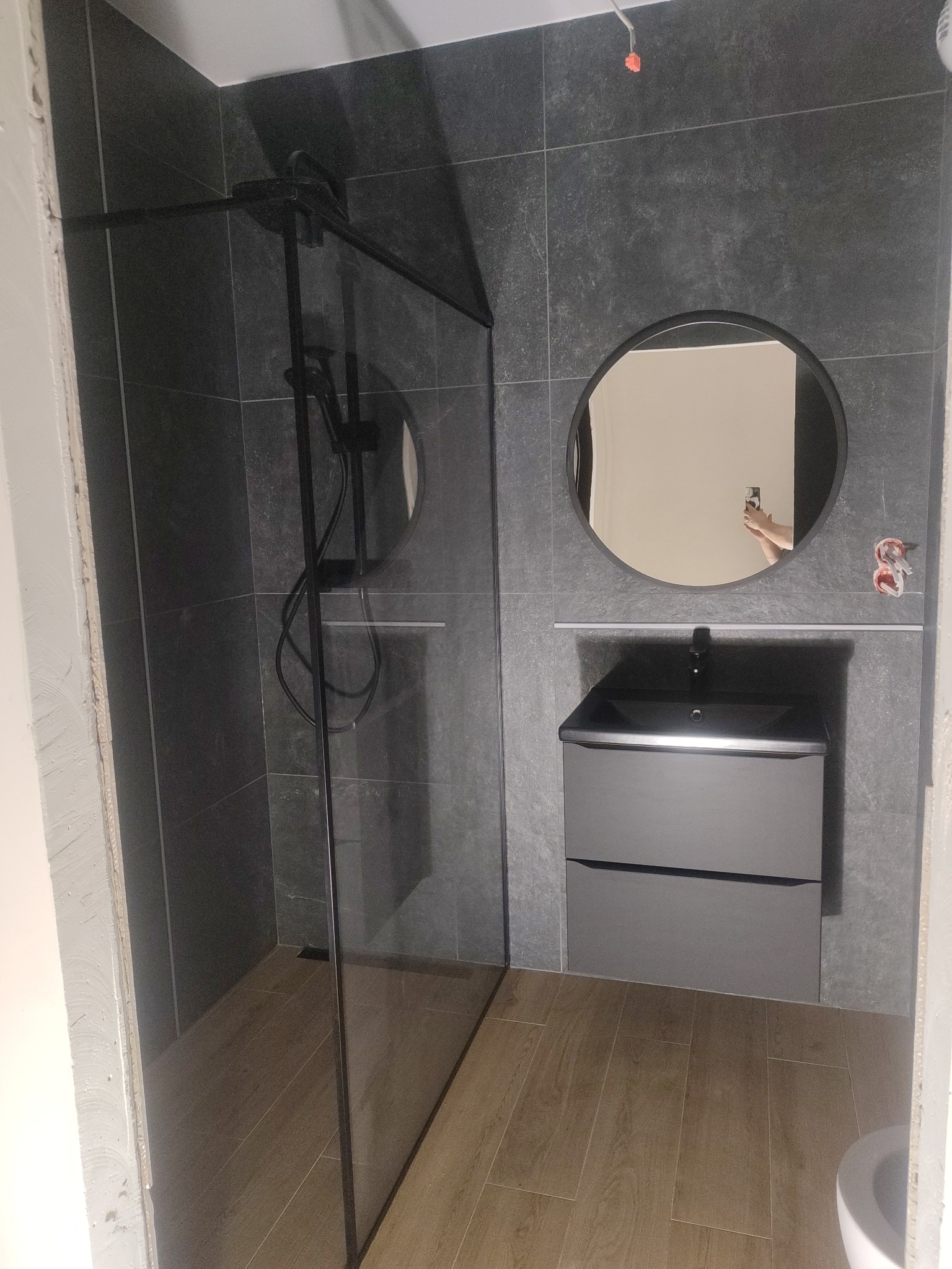 Nowoczesna łazienka w trakcie wykańczania: kabina prysznicowa typu walk-in z czarnymi profilami, umywalka z szafką i okrągłe lustro na grafitowych płytkach.