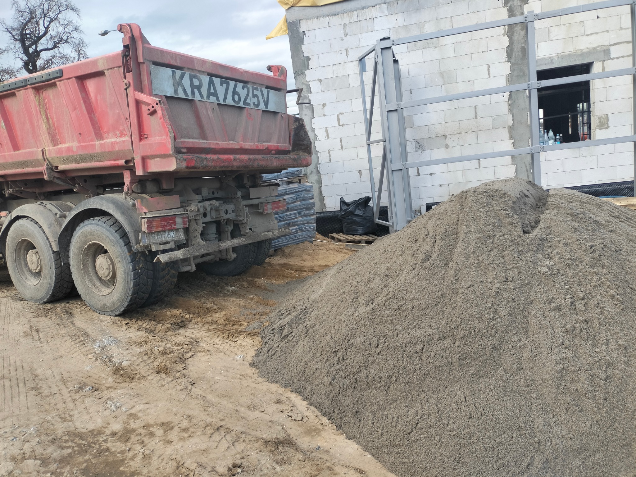 Ciężarówka z numerem KRA7625V obok sterty piasku na placu budowy, widoczny fragment budynku w trakcie wznoszenia z białego bloczka.